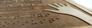 engraved plywood with braille, latin script and the drawing of a hand