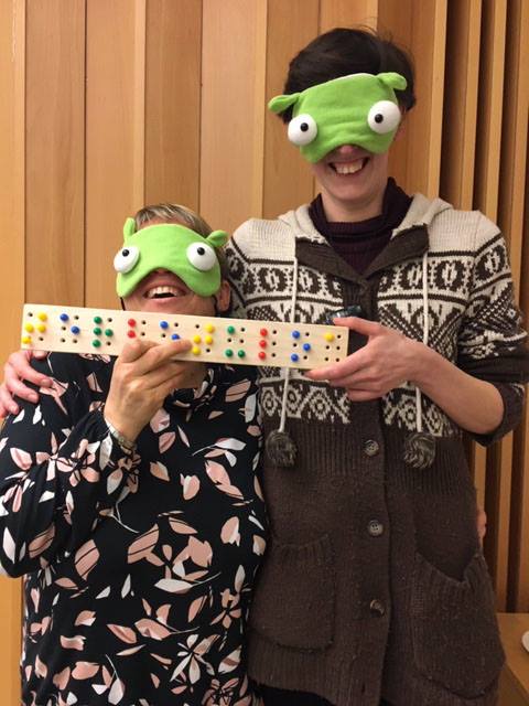 2 persons wearing funny green eyeshades. THey are wearing a wooden board with colored pegs forming the finnish word tervetuloa  (welcome) in braille. 