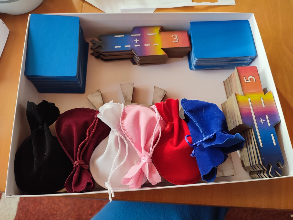 a white box containing 2 stacks of cards, several color pouched and some cardboard scoring tiles